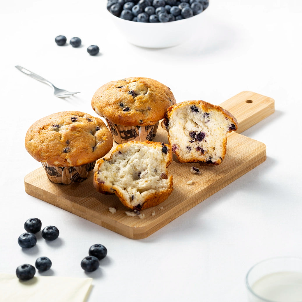 Blueberry muffins on a wooden board with a bowl of blueberries and a glass of milk in the background.