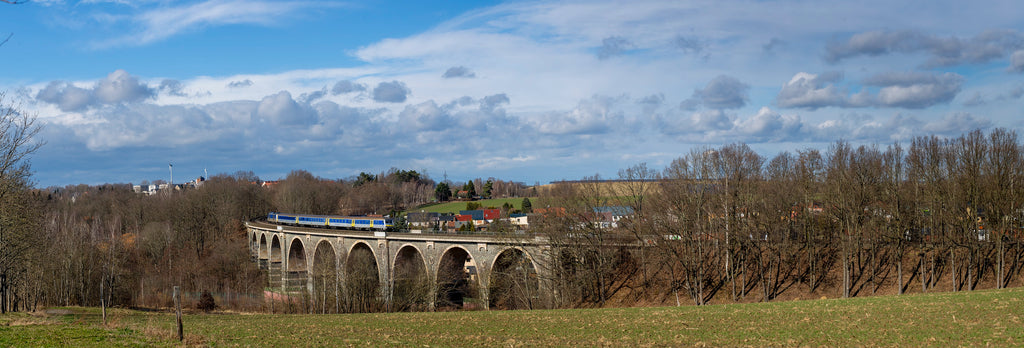 Viaduct with a train passing through, surrounded by trees and fields under a blue sky.
