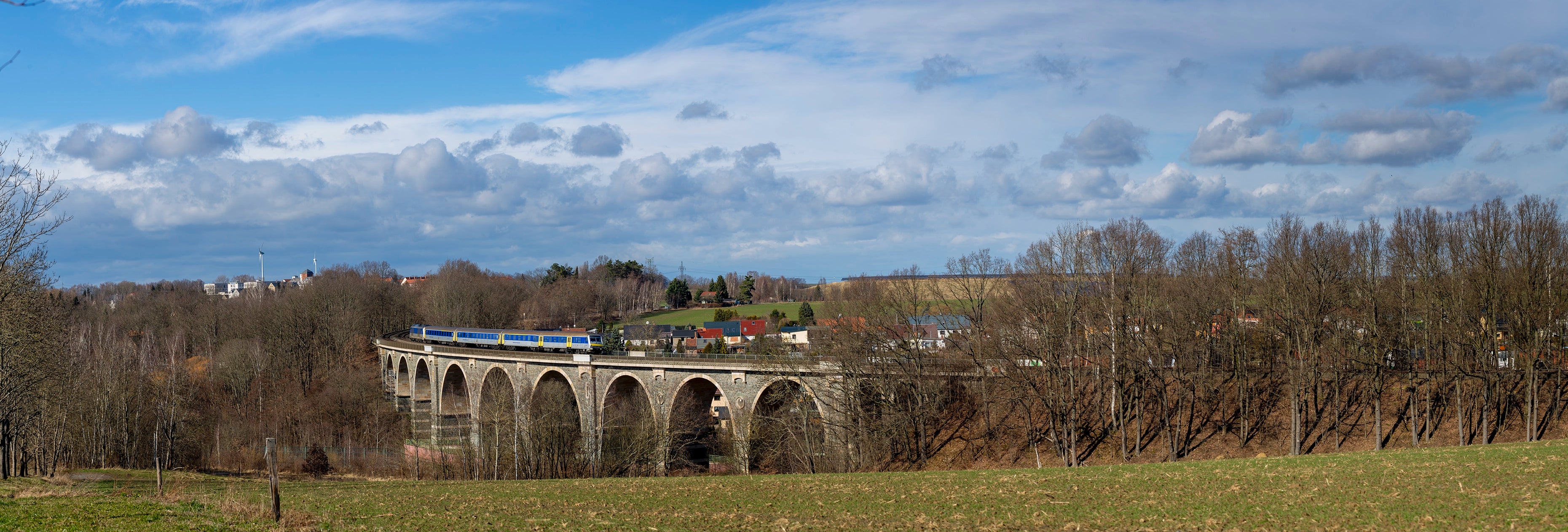 Viaduct with a train passing through, surrounded by trees and fields under a blue sky.