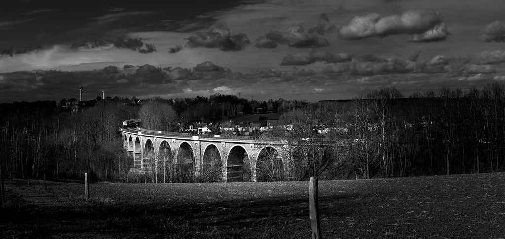 Black and white image of a stone viaduct with trees and clouds in the background