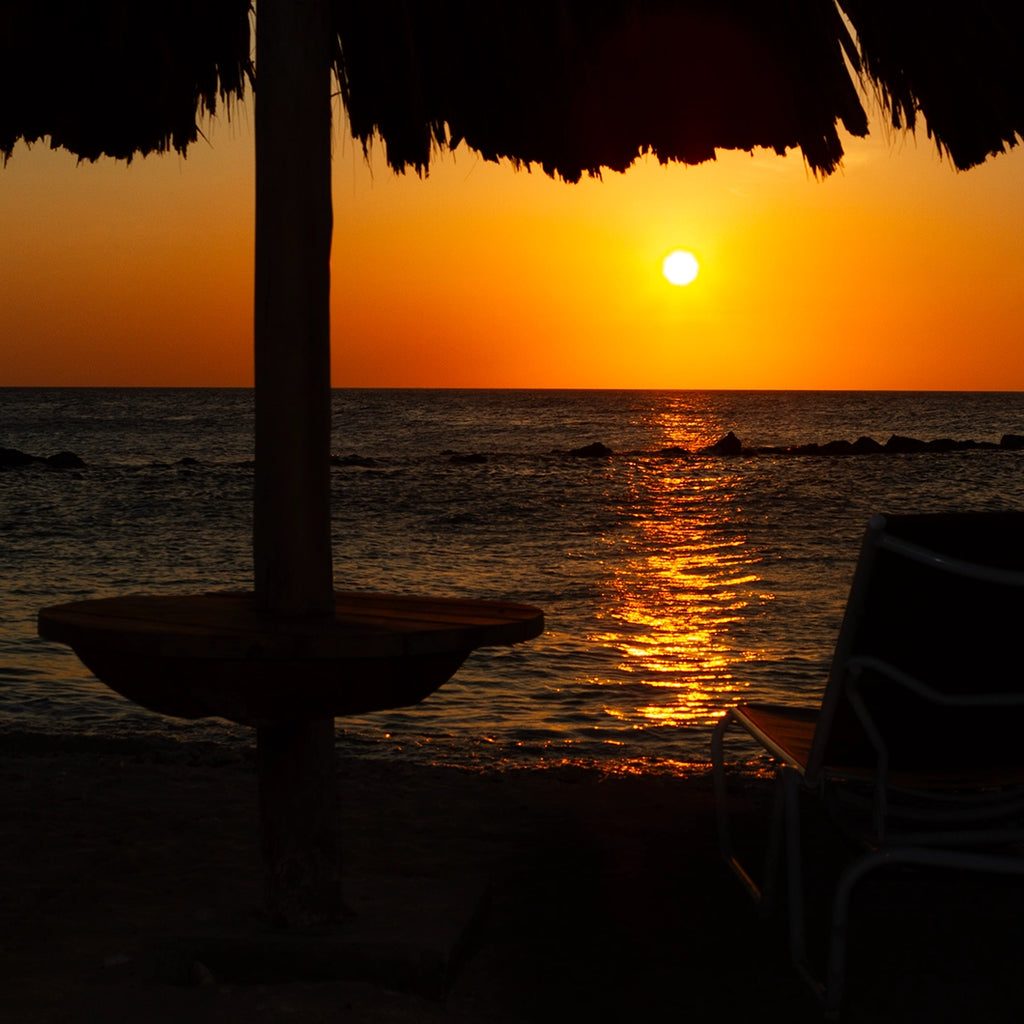 Sunset over the ocean with a thatched-roof structure in the foreground.