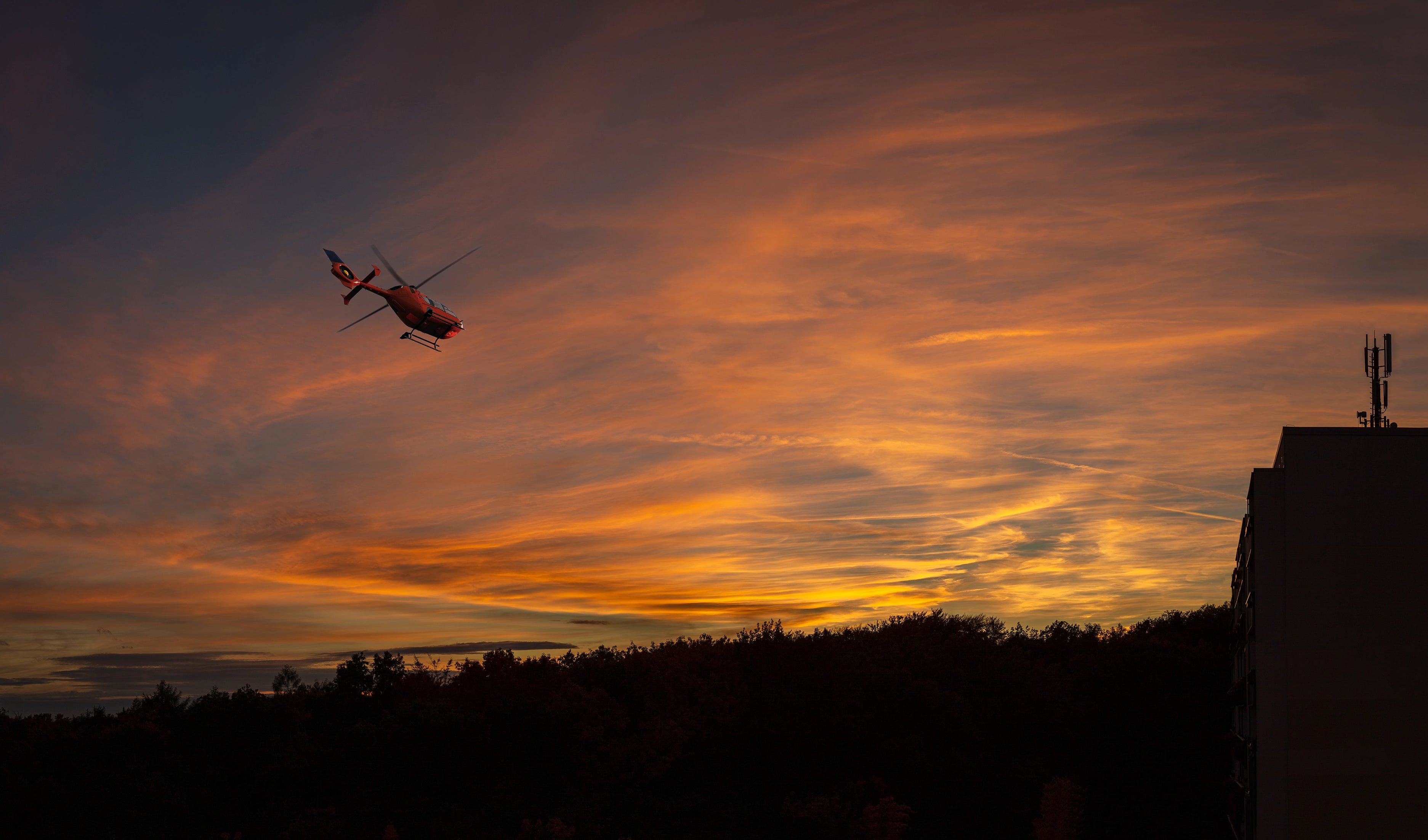Artwork depicting a silhouette of a helicopter in flight against a sunset sky with orange and gold hues.