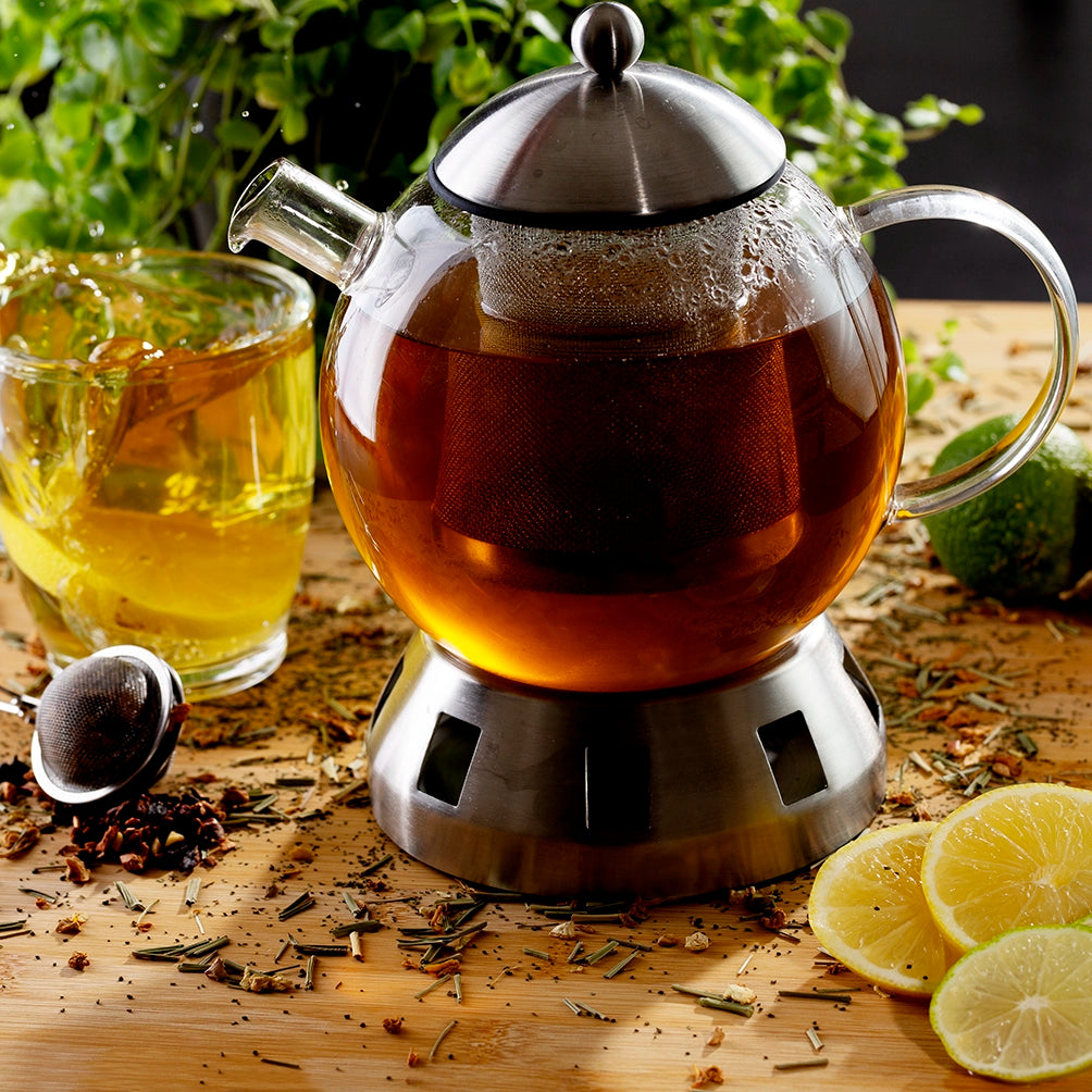 Glass teapot with a metal base on a wooden surface, surrounded by tea leaves and lemon slices.